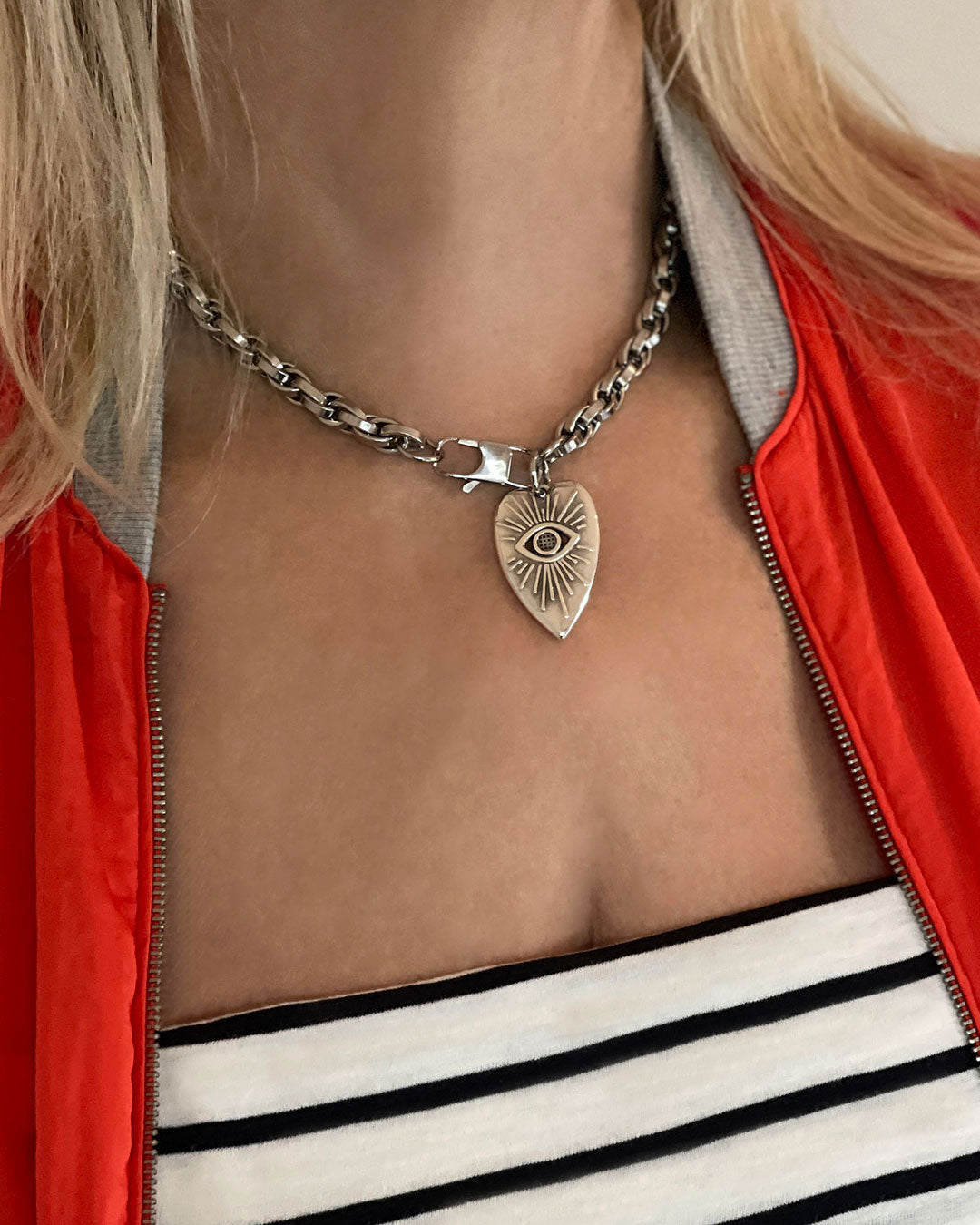 Model in a red jacket and striped t-shirt, wearing a silver chain necklace with a heart-shaped pendant. The heart pendant has a raised eye design with sun rays around the eye.