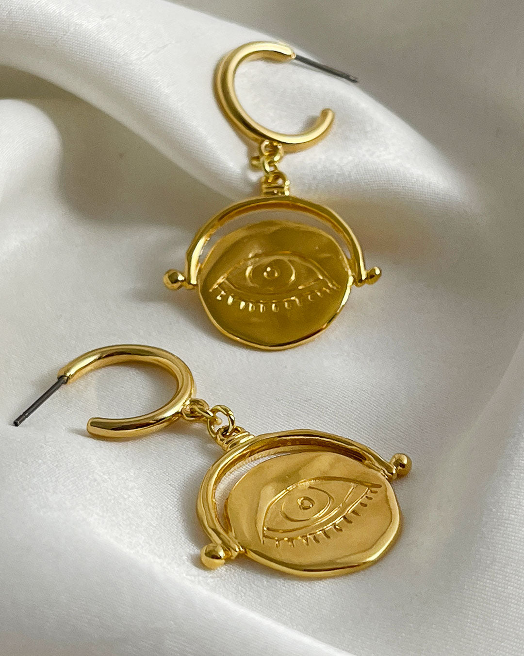 A pair of gold hoop earrings featuring an evil eye round charm on a white satin background