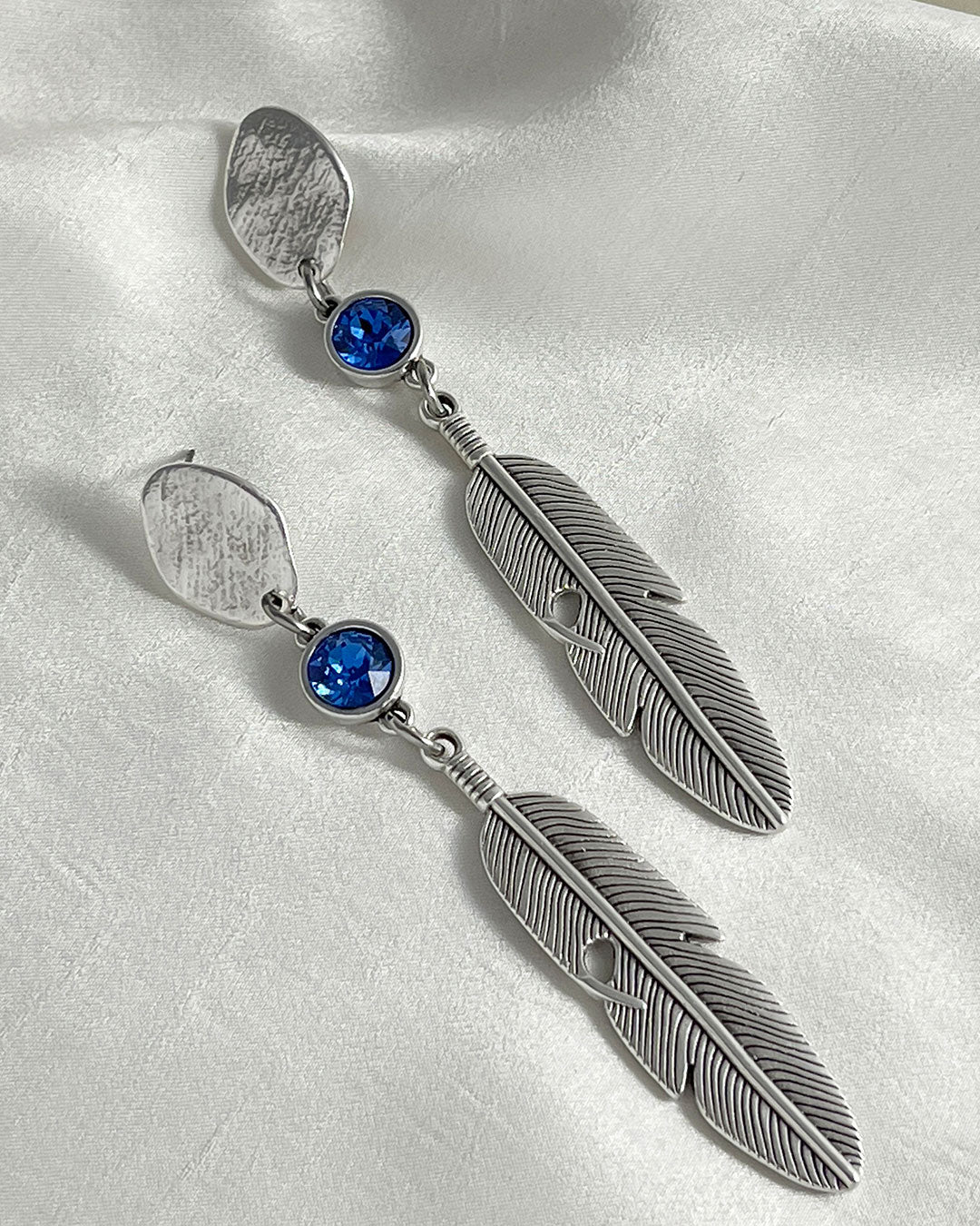 A pair of silver feather earrings with a blue Swarovski crystal. Each earring features a textured silver feather charm dangling from silver oval-shaped studs. The earrings have a bohemian style and are suitable for adding a touch of elegance to any outfit.