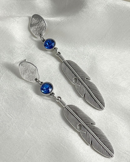 A pair of silver feather earrings with a blue Swarovski crystal. Each earring features a textured silver feather charm dangling from silver oval-shaped studs. The earrings have a bohemian style and are suitable for adding a touch of elegance to any outfit.