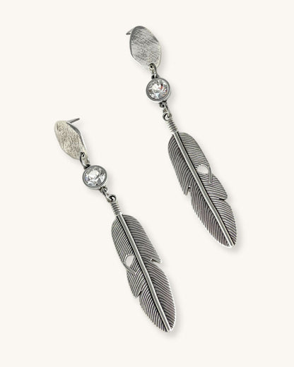 A pair of silver feather earrings with a crystal clear Swarovski. Each earring features a textured silver feather charm dangling from silver oval-shaped studs. The earrings have a bohemian style and are suitable for adding a touch of elegance to any outfit.
