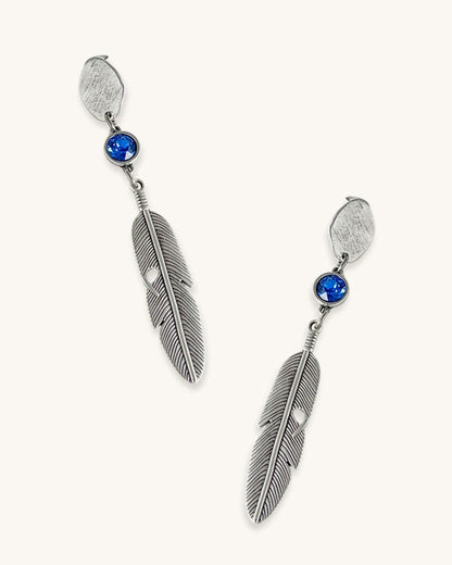 A pair of silver feather earrings with a blue Swarovski crystal. Each earring features a textured silver feather charm dangling from silver oval-shaped studs. The earrings have a bohemian style and are suitable for adding a touch of elegance to any outfit.