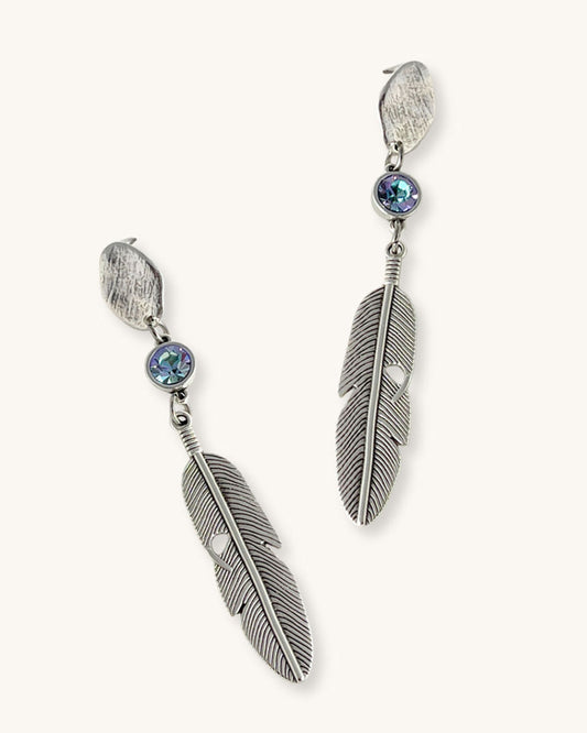 A pair of silver feather earrings with an aquamarine Swarovski crystal. Each earring features a textured silver feather charm dangling from silver oval-shaped studs. The earrings have a bohemian style and are suitable for adding a touch of elegance to any outfit.