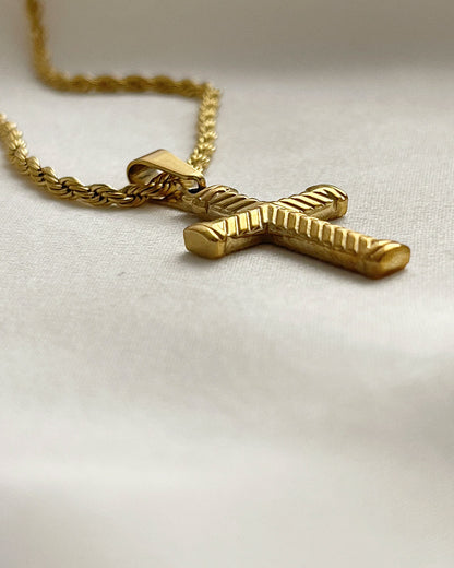 Close up details from a gold rope chain necklace with a gold cross pendant. The cross has a textured design.