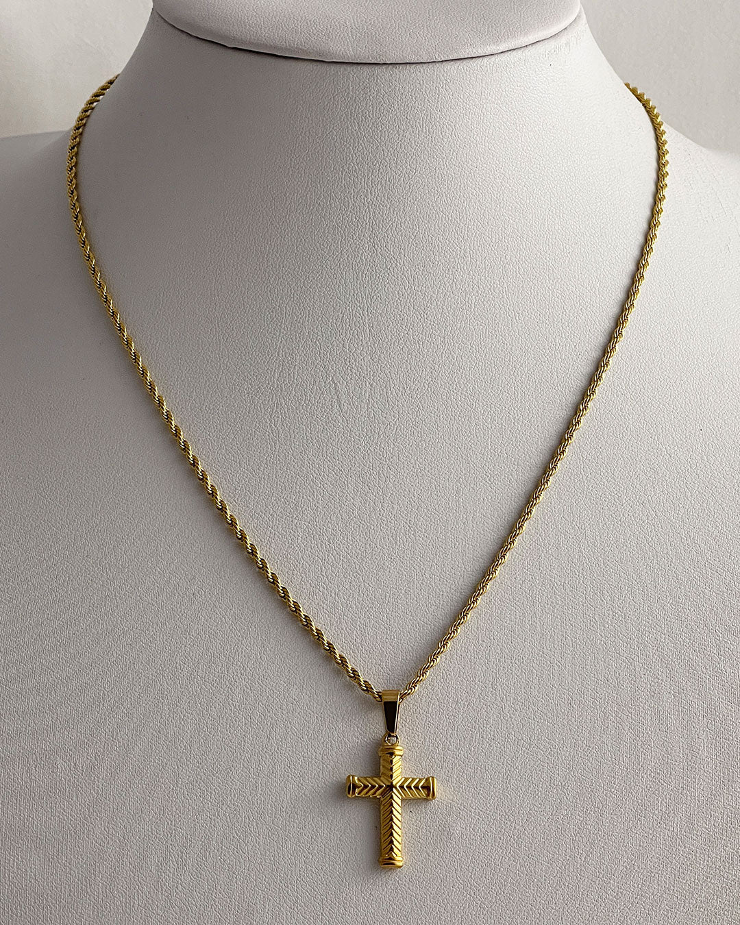 A gold rope chain necklace with a gold cross pendant. The cross has a textured design.