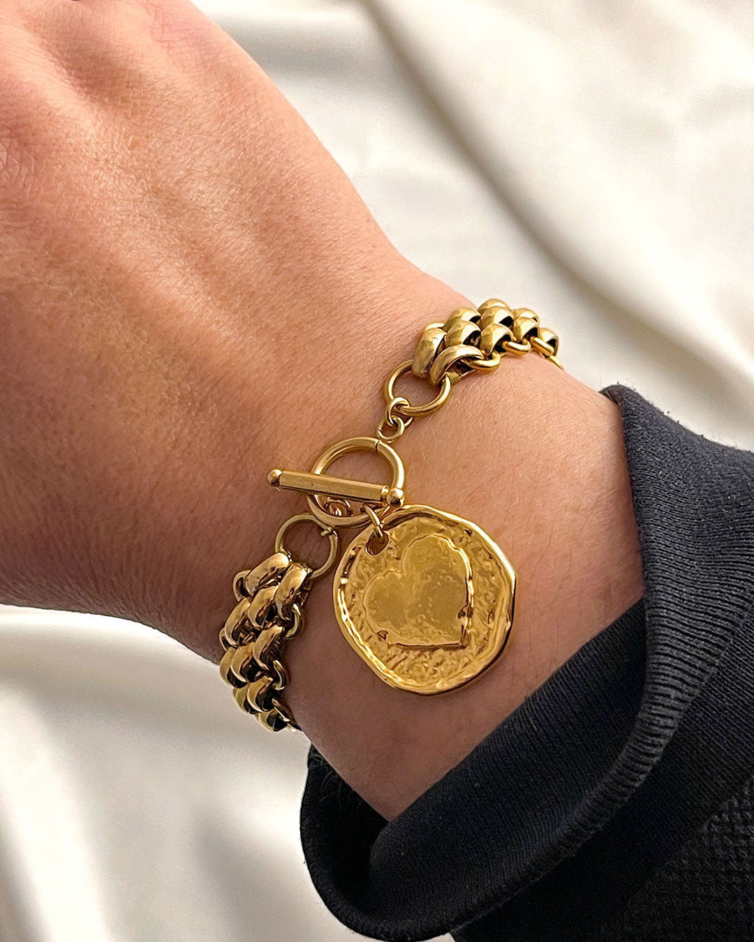 Gold multilink chain bracelet with a heart coin charm on a wrist against a neutral background
