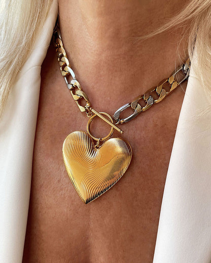 A bold statement chain necklace with a large textured gold heart pendant. The two-tone stainless steel figaro chain blends silver and gold for a striking contrast of luxury.  