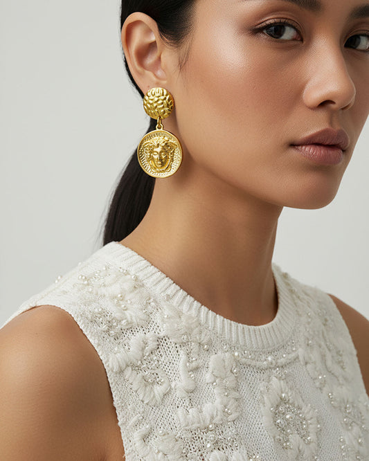 Model wearing a pair of gold earrings featuring a round medusa head charm.
