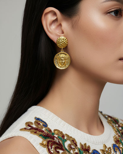 Model wearing a pair of gold earrings featuring a round medusa head charm.