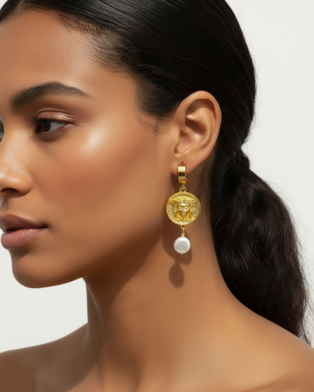 Model wearing a pair of gold hoop earrings adorned with a medusa round charm and a MOP shell pearl