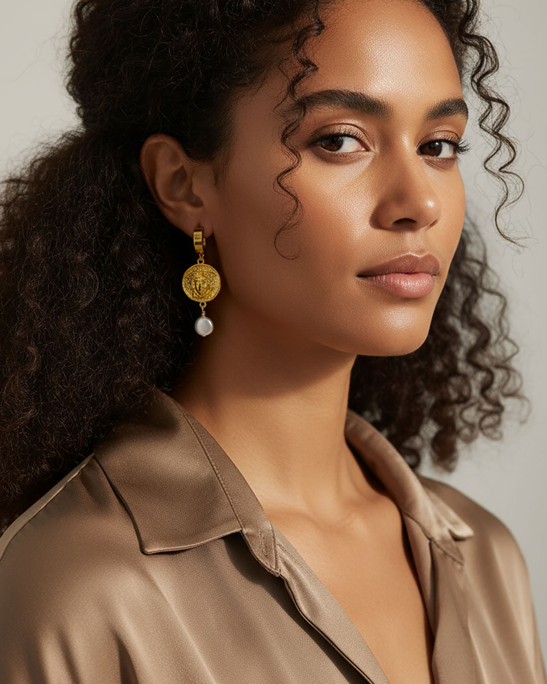 Model wearing a pair of gold hoop earrings adorned with a medusa round charm and a MOP shell pearl