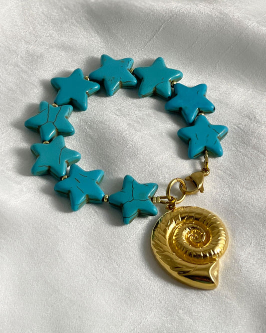 Bracelet with turquoise star-shaped howlite beads and a large gold nautilus shell charm on a white fabric background
