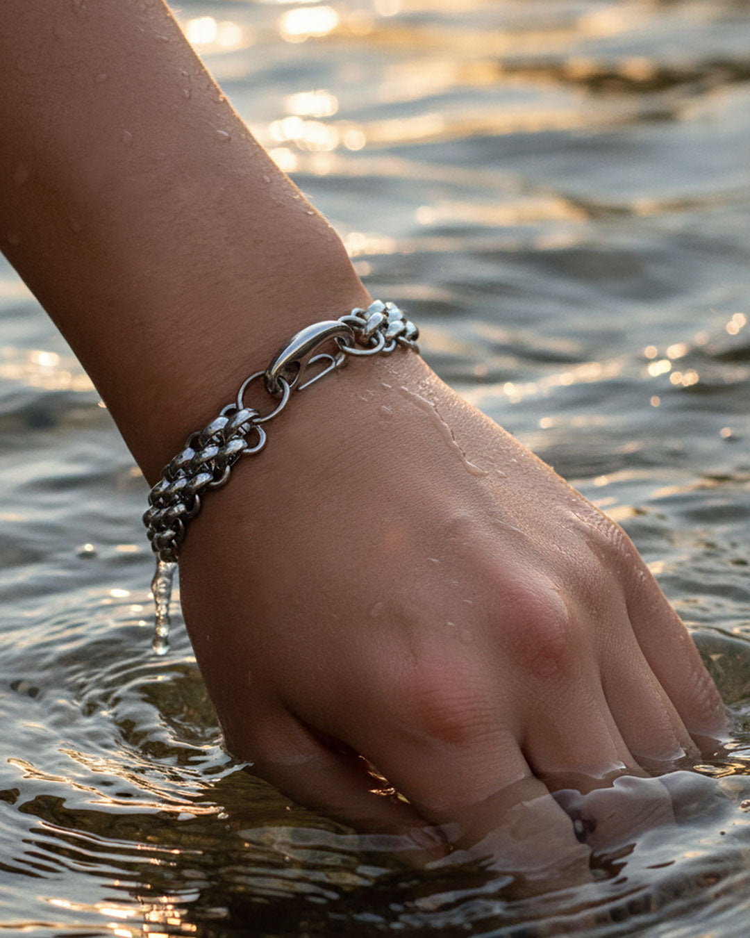 Hand wearing a silver chain bracelet in water with a sunset background