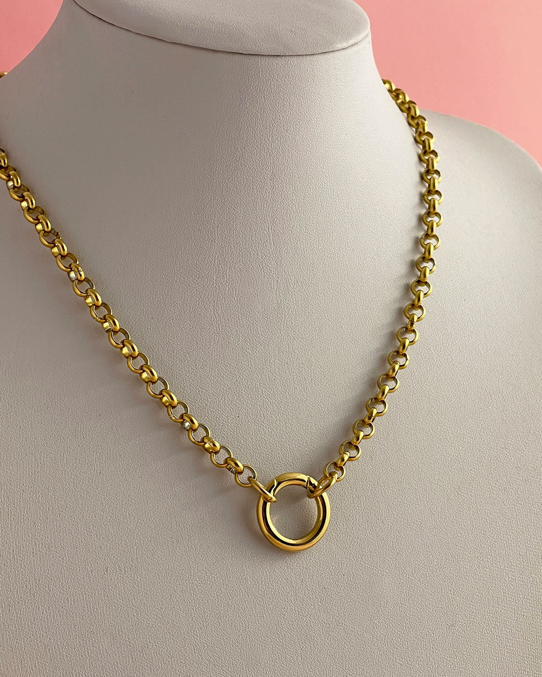 A gold rolo chain necklace with a large circular ring closure, displayed on a white jewelry bust against a soft pink background.