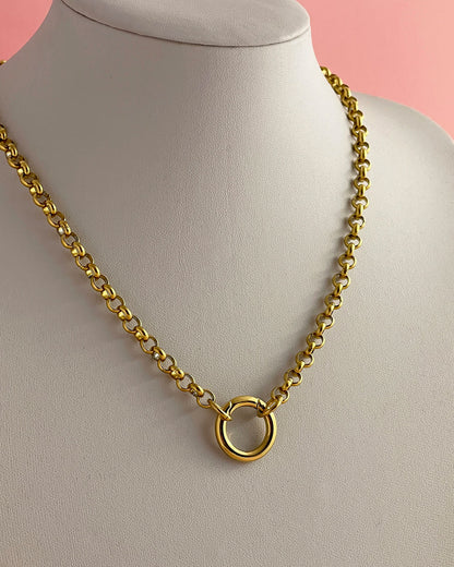 A gold rolo chain necklace with a large circular ring closure, displayed on a white jewelry bust against a soft pink background.