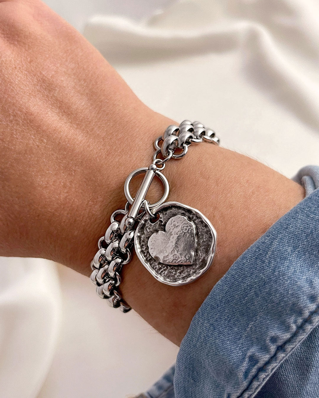 Silver multilink chain bracelet with a heart coin charm on a wrist against a neutral background