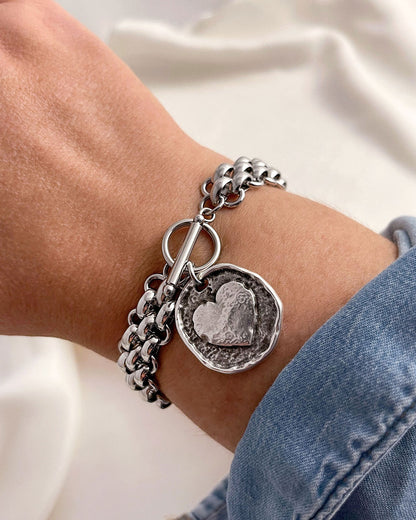 Silver multilink chain bracelet with a heart coin charm on a wrist against a neutral background