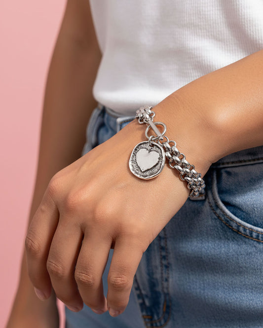 Silver chainmail bracelet with heart charm on a wrist against a pink background