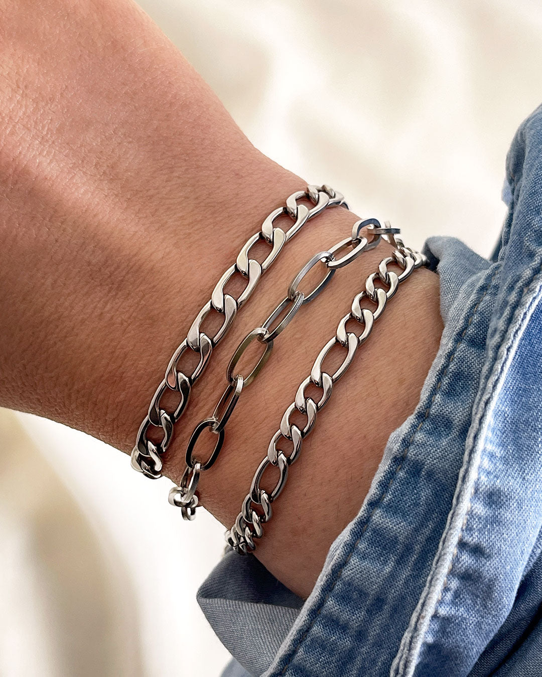 Model wearing three silver chain bracelets with different link styles. The bracelets are all different widths and have a curb chain, paperclip and a figaro chain design.