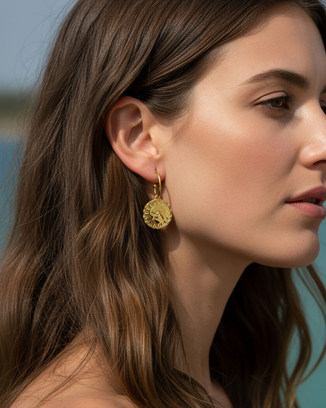 Woman wearing a pair of gold open hoop earrings featuring a round double faced sun moon charm.