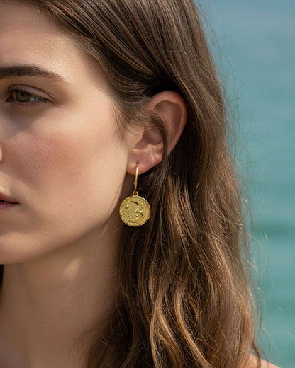 Woman wearing a pair of gold open hoop earrings featuring a round double faced sun moon charm.