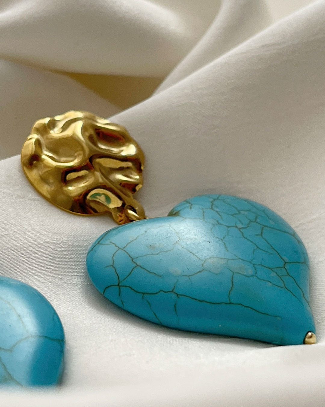 A close-up look at a pair of turquoise howlite heart-shaped earrings. The hearts are hanging from gold hammered studs.
