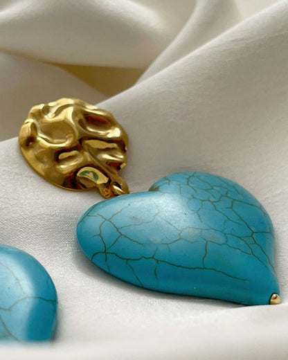 A close-up look at a pair of turquoise howlite heart-shaped earrings. The hearts are hanging from gold hammered studs.