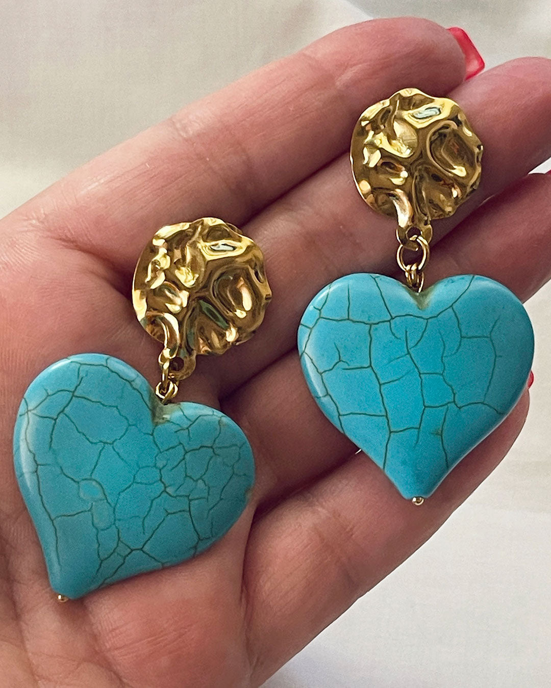 A close-up look at a pair of turquoise howlite heart-shaped earrings. The hearts are hanging from gold hammered studs.