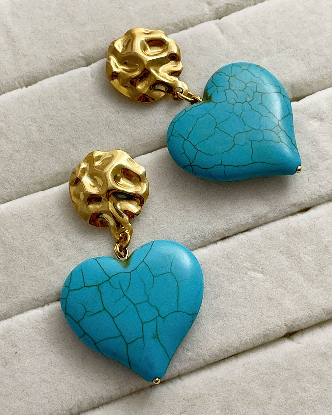 A pair of turquoise howlite heart-shaped earrings. The hearts are hanging from gold hammered studs.