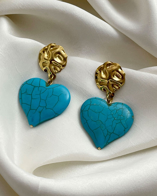 A pair of turquoise howlite heart-shaped earrings. The hearts are hanging from gold hammered studs.