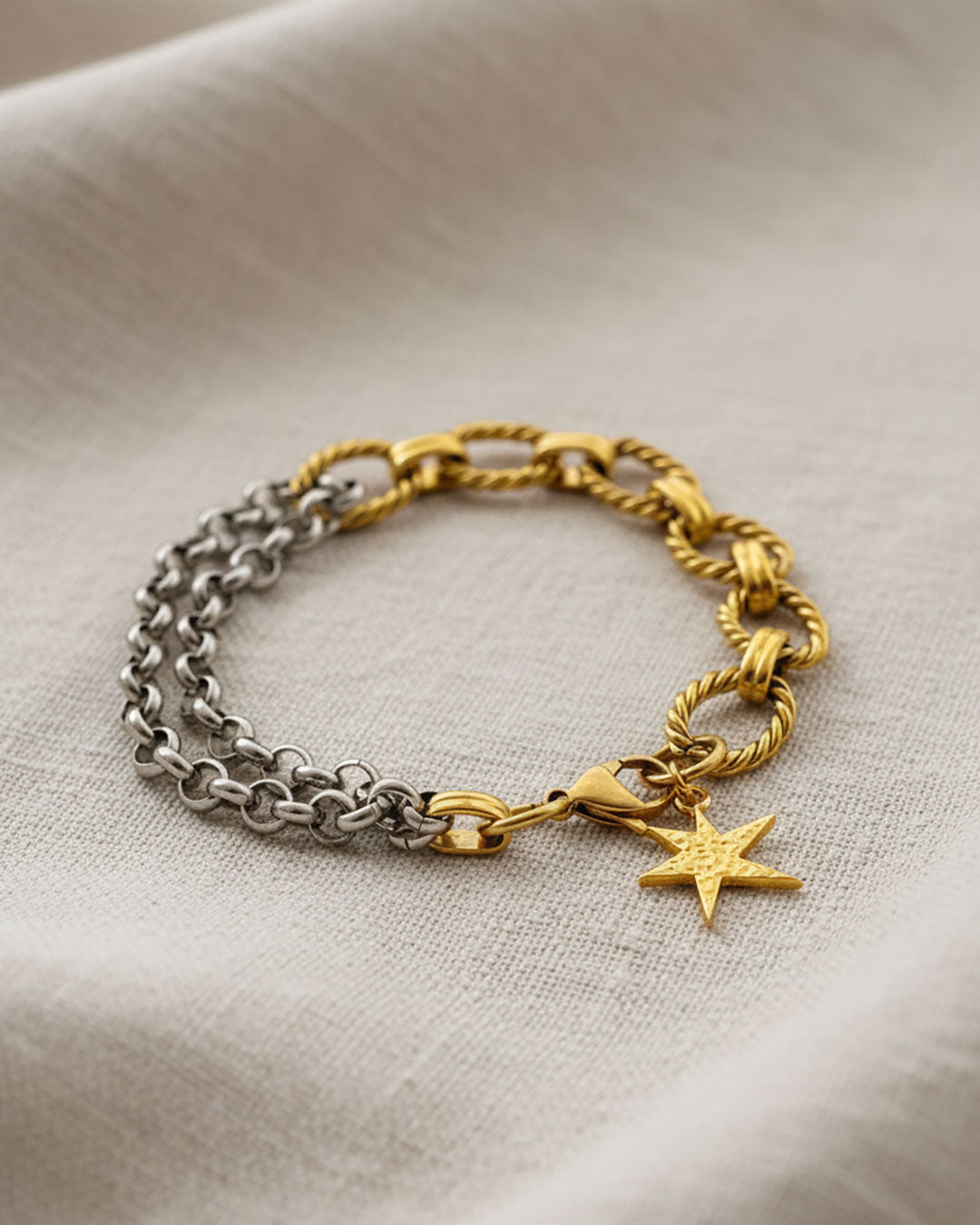 Two chain bracelets, one gold and one silver with a gold charm charm on a fabric background