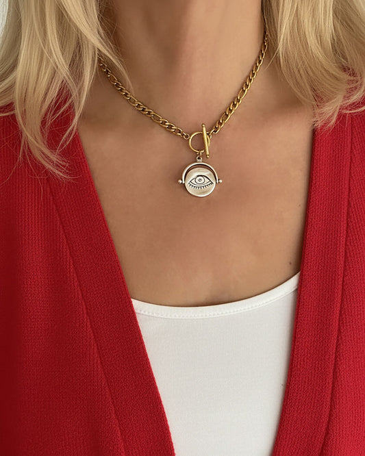 Gold necklace with a silver eye pendant worn by a person in a red cardigan and white top.