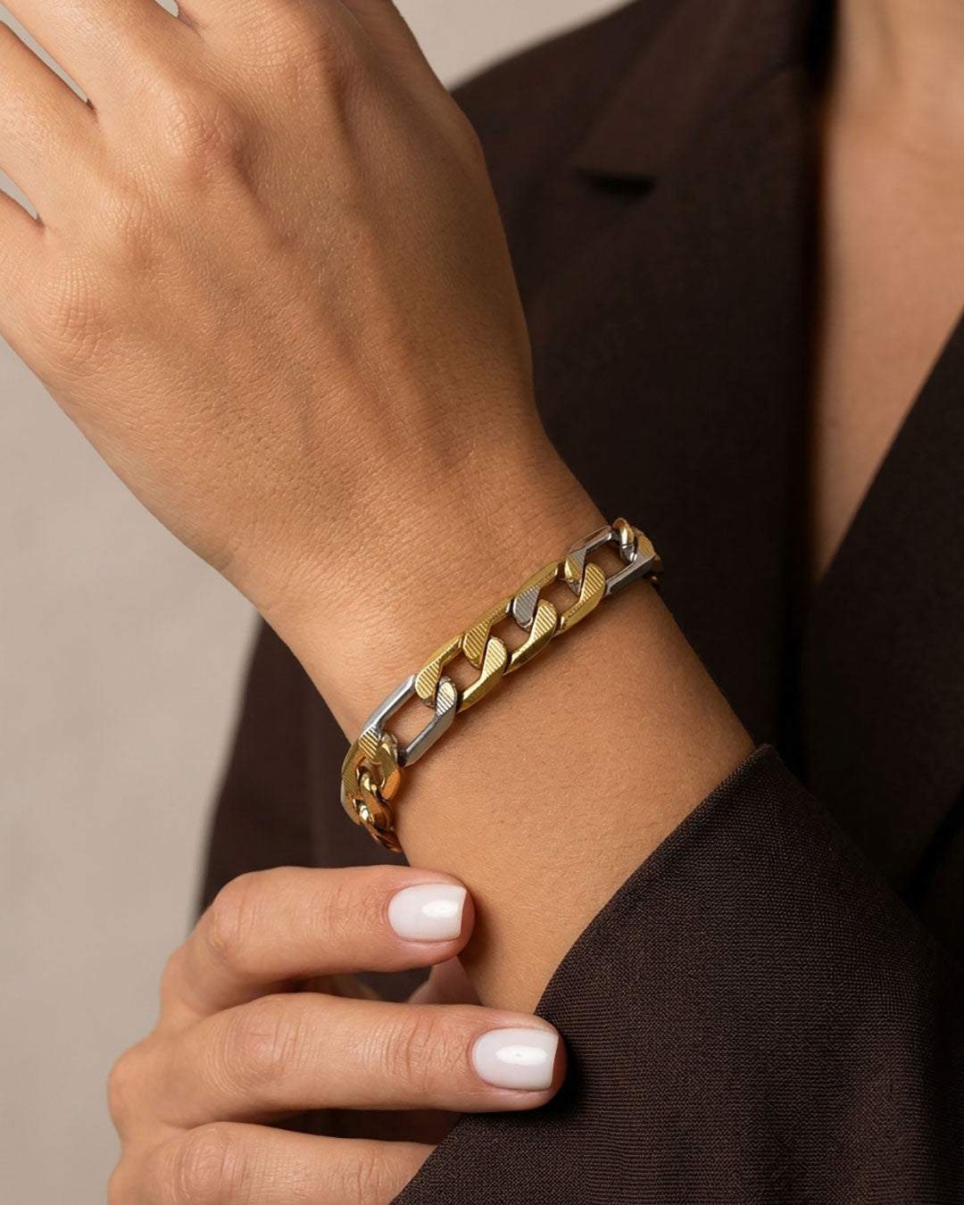 A close up of a woman's wrist wearing a chunky two-tone gold and silver figaro chain bracelet, paired with a dark brown blazer.