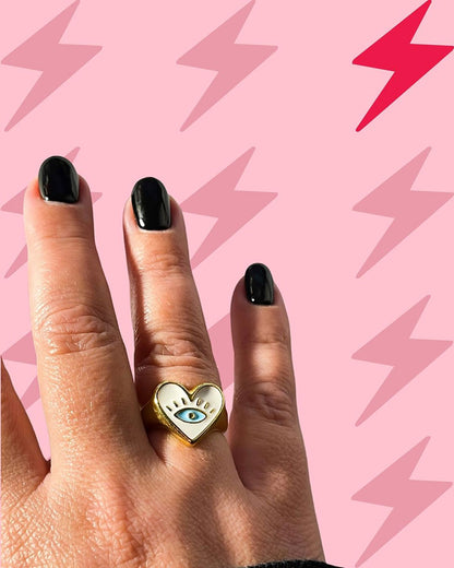 A person's hand displaying a gold chunky heart ring with a white enamel heart and an evil eye design on a pink background with lightning bolts.