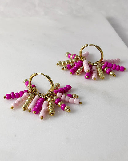 A pair of boho beaded hoop earrings in pink, fuchsia, and gold colors with stainless steel hoops finished in 18K gold, displayed against a white background.