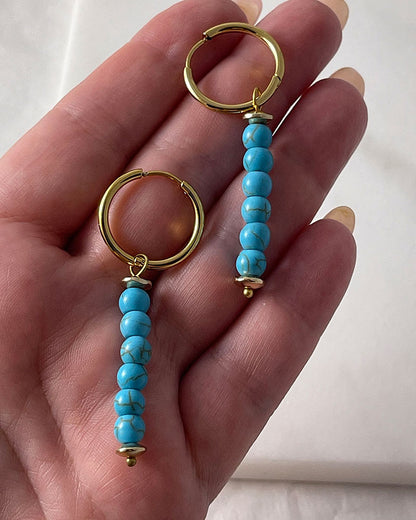 The Turquoise Gem Hoop Earrings, a pair of gold hoop earrings featuring turquoise howlite stones.