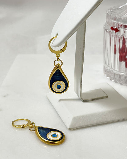 The Eye Drop Earrings, a pair of gold hoop earrings featuring a blue enamel evil eye drop shaped charm.