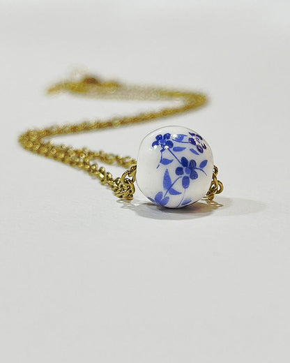 A gold chain necklace with a single white ceramic bead. The bead has a blue floral design.