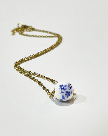 A gold chain necklace with a single white ceramic bead. The bead has a blue floral design.