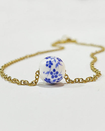 A gold chain necklace with a single white ceramic bead. The bead has a blue floral design.