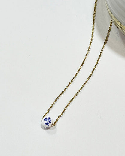 A gold chain necklace with a single white ceramic bead. The bead has a blue floral design.