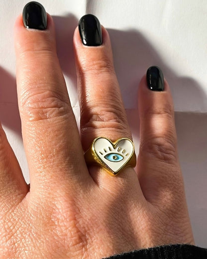 A gold chunky ring with a white enamel heart shape and an evil eye on a finger
