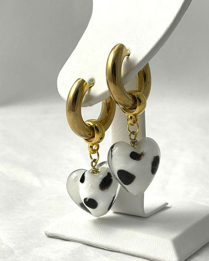 A pair of earrings with gold-plated hoops and white-black glass heart charms displayed on a stand.