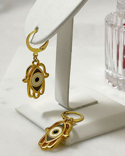 The Hamsa Eye Earrings, a pair of gold hoop earrings featuring a hamsa hand with an eye enamel charm.