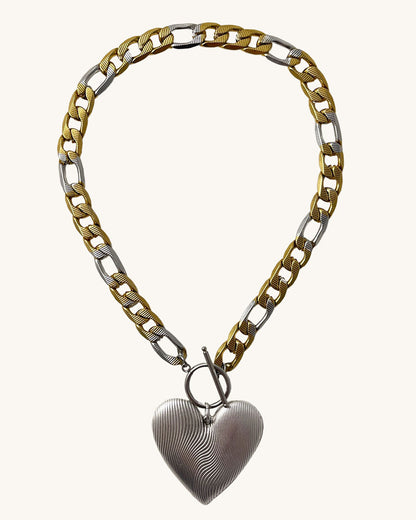 A bold statement chain necklace with a large textured silver heart pendant. The two-tone stainless steel figaro chain blends silver and gold for a striking contrast of luxury.  