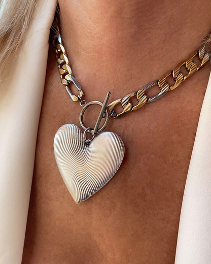 A bold statement chain necklace with a large textured silver heart pendant. The two-tone stainless steel figaro chain blends silver and gold for a striking contrast of luxury.  