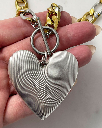 A bold statement chain necklace with a large textured silver heart pendant. The two-tone stainless steel figaro chain blends silver and gold for a striking contrast of luxury.  