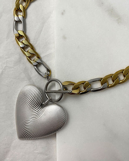 A bold statement chain necklace with a large textured silver heart pendant. The two-tone stainless steel figaro chain blends silver and gold for a striking contrast of luxury.  