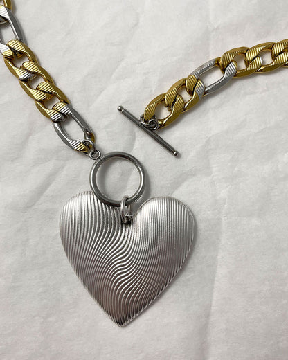A bold statement chain necklace with a large textured silver heart pendant. The two-tone stainless steel figaro chain blends silver and gold for a striking contrast of luxury.  