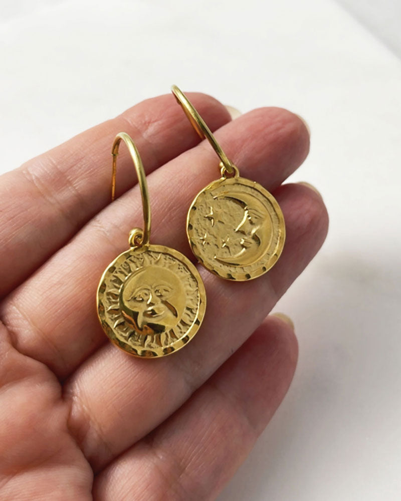 The Sun Moon Hoop Earrings, a pair of gold open hoop earrings featuring a round double faced sun moon charm.